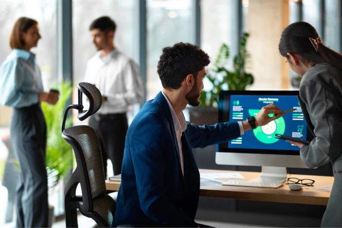 Duas pessoas analisando gráficos de KPIs em um computador, enquanto outros dois colaboradores conversam ao fundo em um escritório moderno.
