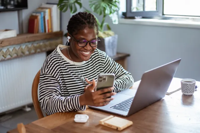 Pessoa sorridente trabalhando remotamente com notebook e celular ao mesmo tempo, em ambiente iluminado e descontraído.