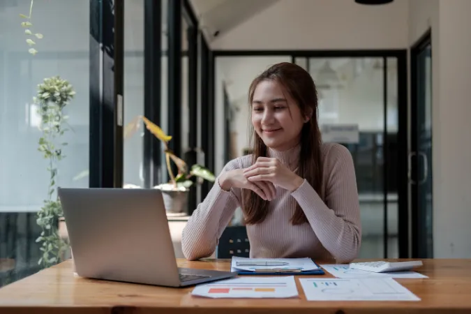 Mulher sorrindo enquanto trabalha no laptop em um escritório iluminado, com documentos e gráficos sobre a mesa, simbolizando o uso eficiente de ERP online.