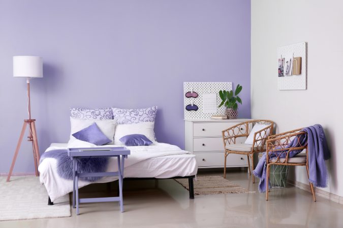 Quarto com parede lilás em destaque, cama com roupas de cama brancas e detalhes em tons de roxo, mesa lateral pintada no mesmo tom da parede, cadeira de vime, luminária de pé e decoração suave, formando um ambiente acolhedor e contemporâneo.