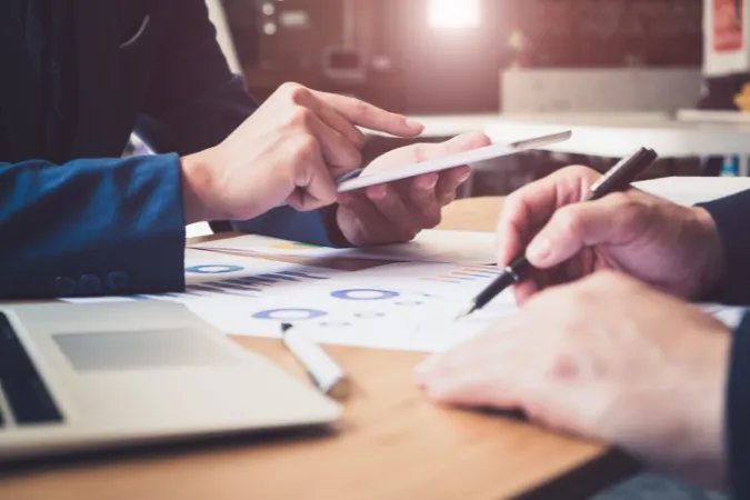 Duas pessoas analisando gráficos e relatórios com auxílio de tablet e laptop sobre uma mesa de trabalho