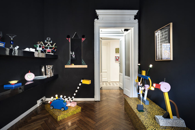 Ambiente interno com paredes pretas e rodapés brancos, decorado com luminárias e objetos escultóricos no estilo Design Memphis. O espaço combina elementos geométricos coloridos, padrões ousados e formas lúdicas sobre pedestais com estampa animal print. Ao fundo, uma porta branca com moldura ornamentada se abre para um corredor claro, criando um contraste entre o estilo clássico da arquitetura e a irreverência do design contemporâneo.