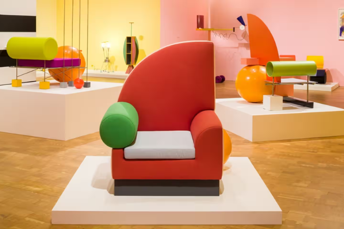 Exposição de peças no estilo Design Memphis, com destaque para uma poltrona vermelha de formas geométricas marcantes, encosto semicircular e apoio de braço cilíndrico verde. Ao fundo, outras mobílias coloridas com volumes esculturais, ambientadas em um espaço com paredes rosa e amarela, piso de madeira e iluminação suave que valoriza o caráter artístico e experimental do mobiliário.