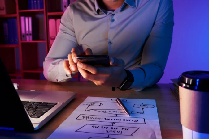 Pessoa analisando fluxograma de projeto de software em ambiente de trabalho com iluminação neon, enquanto segura o celular.