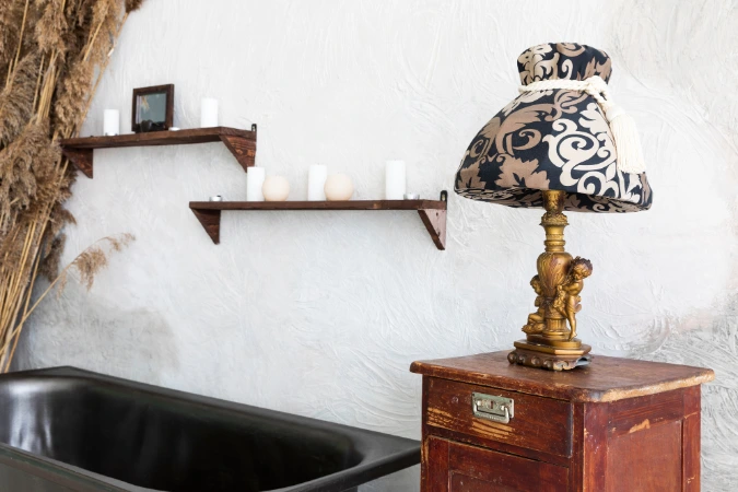 Banheiro com decoração industrial, incluindo banheira preta, parede texturizada clara, prateleiras com velas e móvel antigo com luminária decorativa.