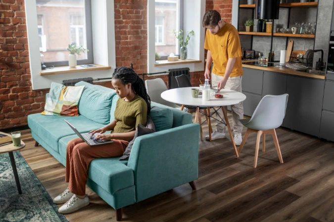Casal em apartamento pequeno com sofá, mesa de jantar e cozinha integrados no mesmo ambiente.