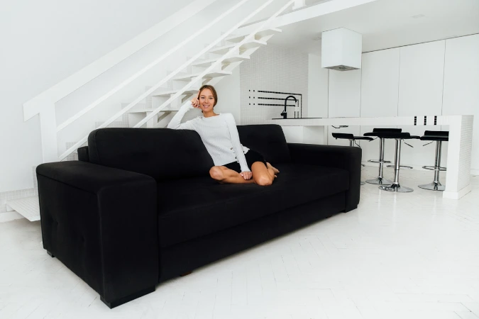 Mulher sentada em sofá preto em loft minimalista com escada e cozinha integrada.