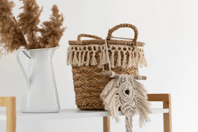 Detalhe de cesta de palha com franjas decorativas e arranjo de flores secas em jarra branca sobre prateleira.
