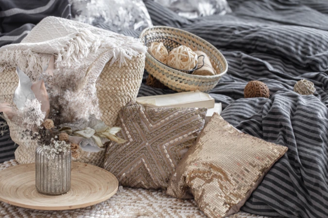 Almofadas decorativas com brilho, cestos artesanais e flores secas sobre cama com roupa de cama listrada em tons sóbrios.