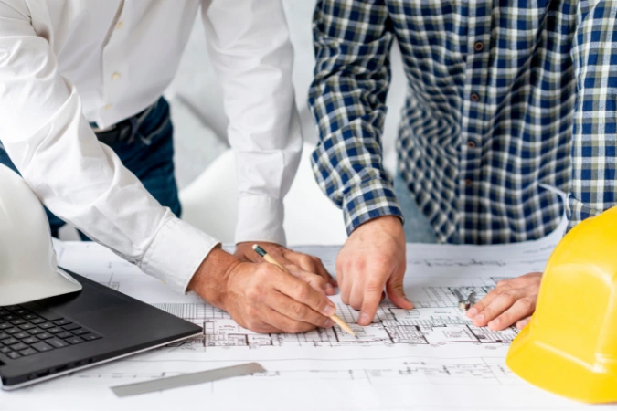 Dois profissionais analisando juntos uma planta baixa sobre a mesa, com capacetes de segurança próximos e um notebook ao lado.