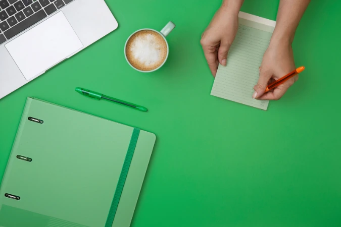 Mesa de trabalho com fundo verde, caderno, canetas, café e notebook organizados de forma minimalista
