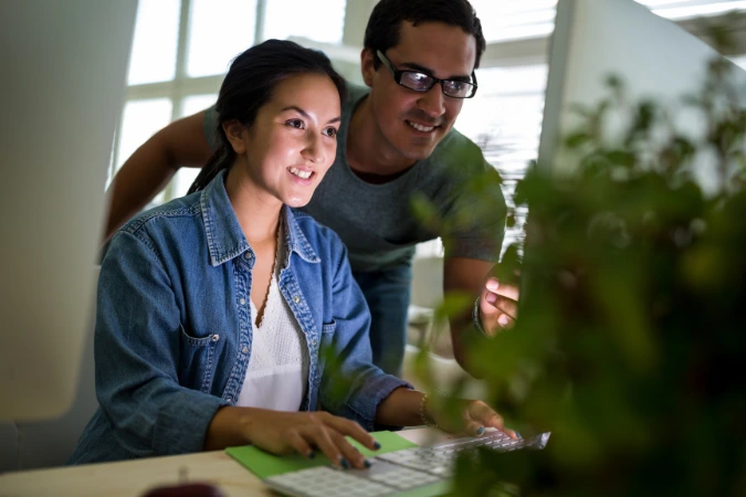 Duas pessoas trabalhando juntas em frente ao computador, com plantas no primeiro plano e iluminação natural ao fundo