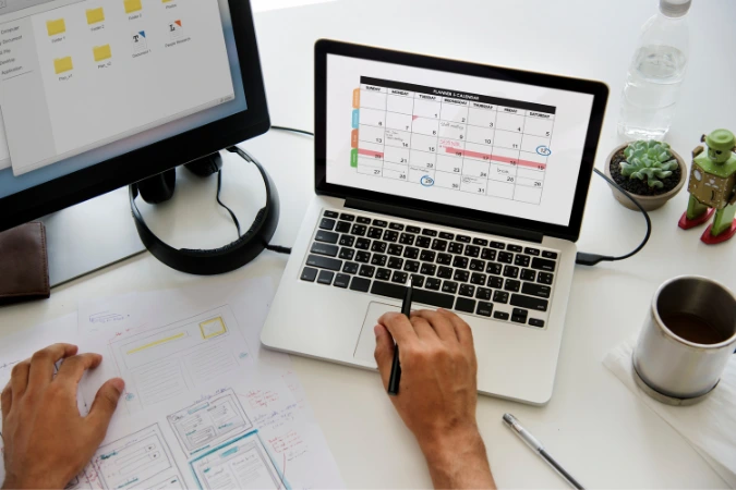 Pessoa trabalhando com calendário digital aberto no notebook ao lado de planilhas, anotações e tela auxiliar com pastas de projeto.