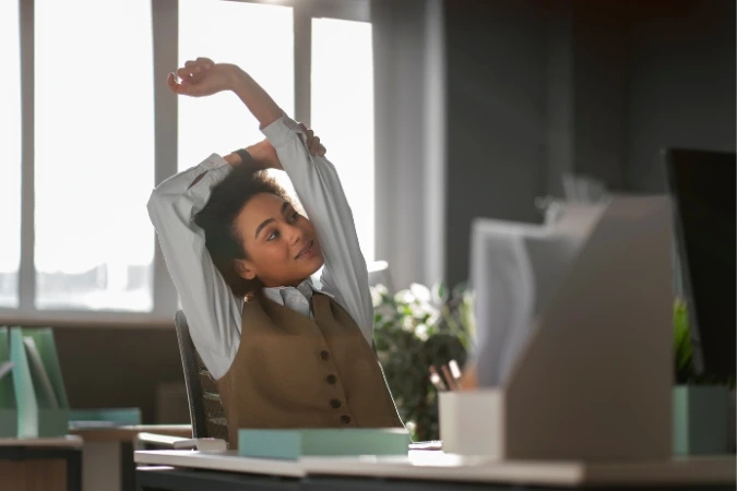 Mulher jovem se espreguiçando em frente ao computador em um escritório iluminado, demonstrando alívio e foco no bem-estar no trabalho.
