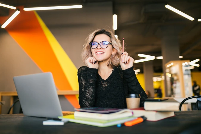 Profissional jovem sorrindo em frente ao notebook, cercada de cadernos e livros em um espaço de trabalho moderno e criativo.