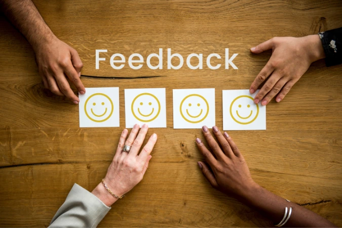 Quatro mãos segurando cartões com rostos sorridentes ao redor da palavra feedback sobre uma mesa de madeira