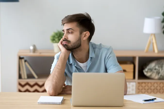 Homem sentado em frente ao notebook, com expressão de tédio e olhar perdido, representando ociosidade no trabalho remoto por falta de organização ou clareza de tarefas.