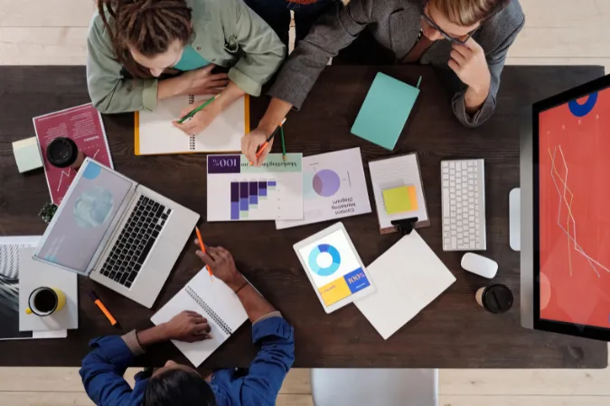 Profissionais reunidos em mesa de trabalho com gráficos, tablets e notebooks, organizando tarefas e discutindo entregas de projeto.
