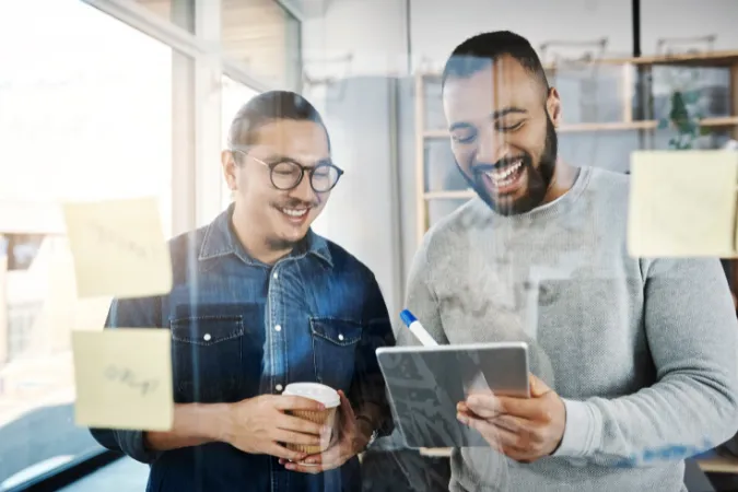 Dois profissionais sorrindo enquanto analisam um tablet em frente a um painel com post-its