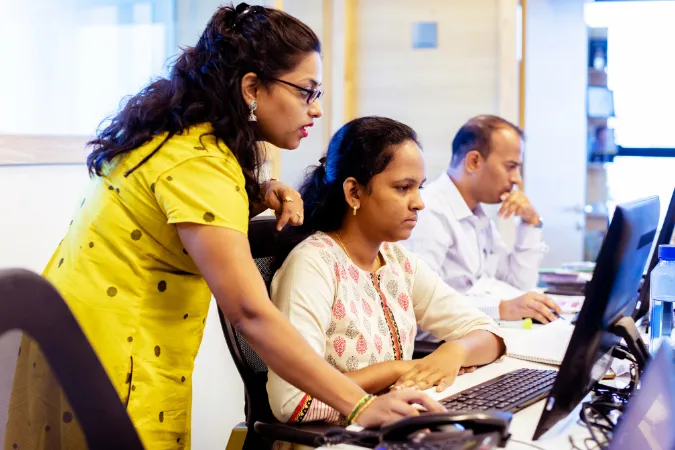 Duas mulheres analisando informações no computador com outro colega ao fundo concentrado