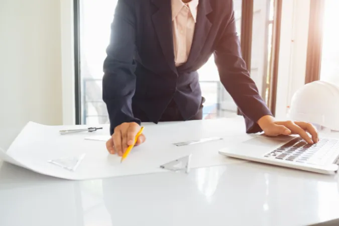 Pessoa de terno analisando planta arquitetônica sobre a mesa enquanto opera um notebook
