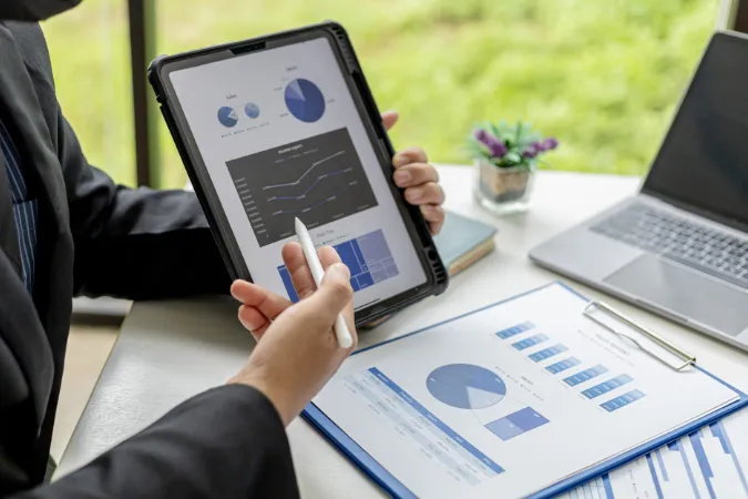Pessoa segurando tablet com gráficos e relatórios de vendas, com planilhas sobre a mesa.