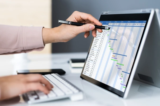Tela de computador exibindo gráfico de Gantt sendo usado em ambiente corporativo, com mão apontando para tarefas
