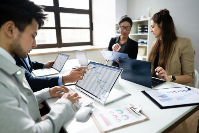 Equipe técnica reunida em torno de computadores com gráficos e cronogramas, representando a integração do cloud MRP com sistemas ERP em ambientes de engenharia
